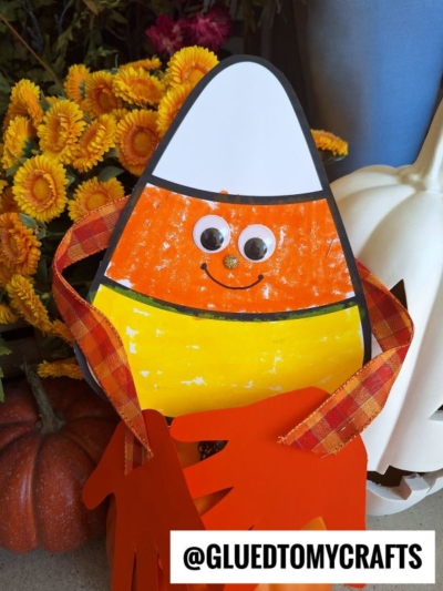 A handmade candy corn hug craft with googly eyes, an orange pom-pom nose, and plaid ribbon arms, displayed in front of fall flowers, a pumpkin, and the handle @GLUETOMYCRAFTS at the bottom.
