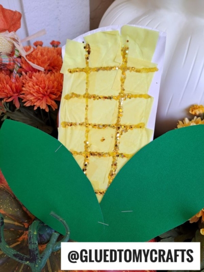 A handmade corn craft with yellow tissue paper and gold glitter on a white paper plate, decorated with two large green paper leaves at the bottom. Orange flowers and pumpkins are in the background.