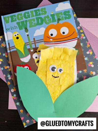 A children’s book titled “Veggies with Wedgies” is paired with a cute paper plate corn craft made of yellow tissue paper, green leaves, and googly eyes on top. @GLUEDTOMYCRAFTS is written at the bottom.