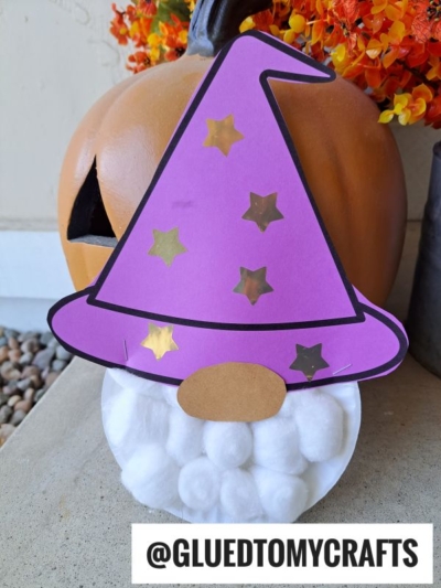 A paper plate wizard gnome with a purple hat decorated with gold stars, a cotton ball beard, and an oval nose sits in front of a carved pumpkin and orange flowers. Text at the bottom reads @GLUEDTOMYCRAFTS.