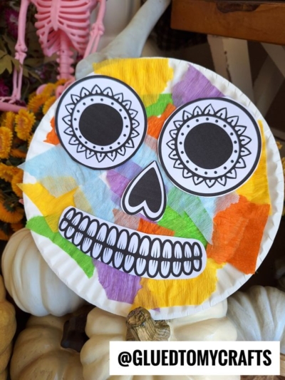 A vibrant paper plate sugar skull decorated with black and white eyes, nose, and mouth, plus bright tissue paper accents. Pumpkins and a pink skeleton sit in the background. Text: @GLUETOMYCRAFTS.