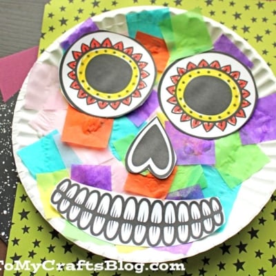 A colorful paper plate sugar skull craft, decorated with tissue paper squares and paper cutouts for the eyes, nose, and mouth.