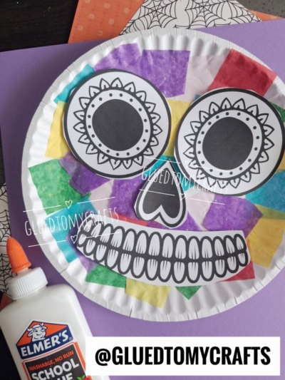 A colorful Day of the Dead paper plate sugar skull craft featuring a vibrant skull face, black and white facial features, tissue paper squares, Elmers glue, and purple paper. @GLUEDTOMYCRAFTS is displayed at the bottom.