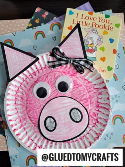 A pink paper plate pig craft with large googly eyes, black-tipped ears, a snout, and a plaid bow—perfect for fork painting crafts—sits on rainbow-patterned paper next to the book I Love You, Little Pookie.