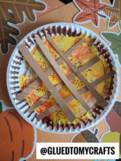 A paint splat pie paper plate craft decorated to look like a pumpkin pie with orange and yellow paint and brown paper strips as crust, placed on a fall-themed background with leaves and pumpkins.