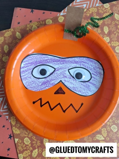 An orange paper plate decorated as a superhero pumpkin Halloween mask with googly eyes, a purple crayon mask, a zigzag mouth, and a small triangular nose. The plate is set on autumn-themed paper with a green pipe cleaner.