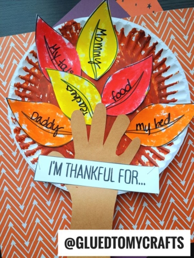 A Thanksgiving craft shows a hand-shaped cutout as a tree trunk with paper leaves listing things to be thankful for, like Mommy, food, daddy, and my bed. This thankful tree craft captures gratitude in a creative way. @GLUEDTOMYCRAFTS is written at the bottom.