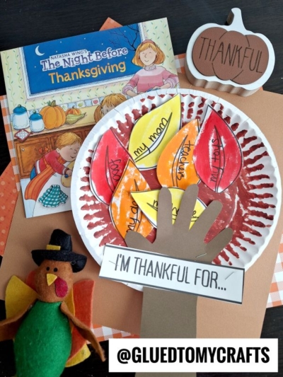 A Thanksgiving craft scene with a book, a paper plate turkey featuring colored paper feathers labeled with things to be thankful for, a handprint cutout, a paper pumpkin, a plush turkey, and an adorable thankful tree craft. Tag: @gluedtomycrafts.