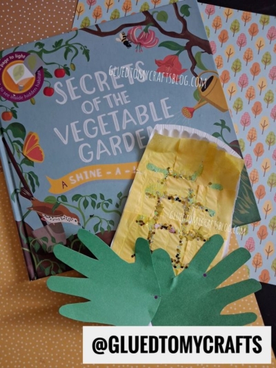Book titled Secrets of the Vegetable Garden featuring a charming corn paper plate craft with green paper leaves and an Instagram handle @GLUEDTOMYCRAFTS, all displayed on a cheerful polka dot surface.