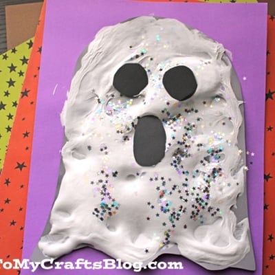 A puffy paint ghost made from white fluffy material, black paper cutouts for eyes and mouth, and sparkly star-shaped glitter sits on purple paper. Colorful star-patterned sheets are beneath it.
