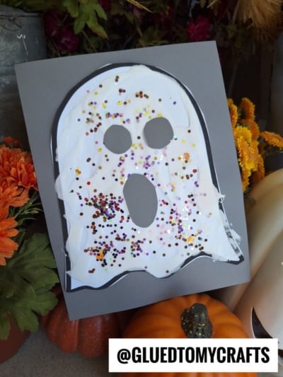 A puffy paint ghost made from white paint and glitter on black paper is displayed among pumpkins and orange flowers. The ghost has eye and mouth holes cut out. @GLUEDTOMYCRAFTS is written at the bottom.