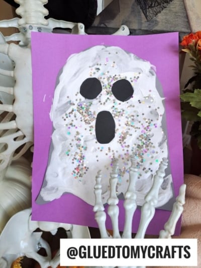 A child’s puffy paint ghost craft made with white paint, black paper eyes and mouth, and colorful star glitter on purple paper. A skeleton prop holds the artwork. Text reads @GLUEDTOMYCRAFTS.