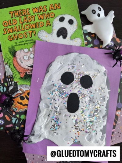 A children’s book titled There Was an Old Lady Who Swallowed a Ghost!, a ghost figurine, plastic spiders, and a handmade puffy paint ghost craft with white paint and glitter on purple paper are arranged on a patterned surface.