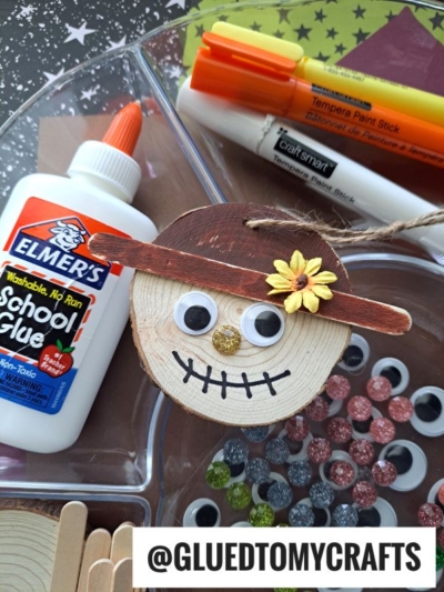 A charming wood slice scarecrow craft with googly eyes, a flower, and a marker smile is displayed, surrounded by glue, craft sticks, markers, glitter glue, and extra googly eyes. This adorable scarecrow ornament project is perfect for creative afternoons. @GLUEDTOMYCRAFTS text is at the bottom.