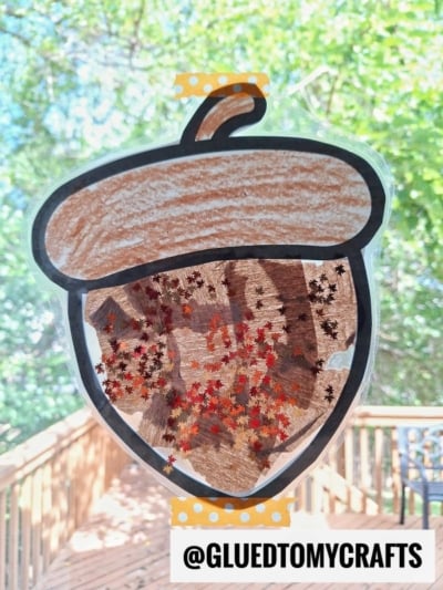 A handmade acorn suncatcher is taped to a window. The acorn is colored brown and filled with tiny red, orange, and brown leaf-shaped confetti. Trees and a wooden deck are visible outside. Text reads @gluedtomycrafts.