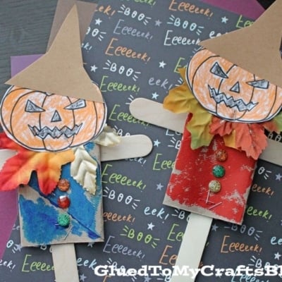 Two handmade toilet paper roll scarecrows with colored pumpkin faces, popsicle stick arms, and decorated shirts are displayed on Halloween-themed paper and surrounded by colored construction paper.