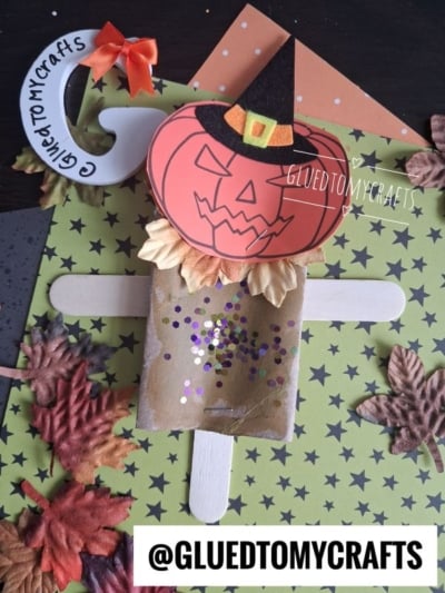 Pumpkin scarecrow craft made from paper, felt, a cardboard tube, glitter, popsicle sticks, and fake leaves on a green star-patterned background. This charming toilet paper roll scarecrow is perfect for fall crafting fun. @gluedtomycrafts is written on the image.