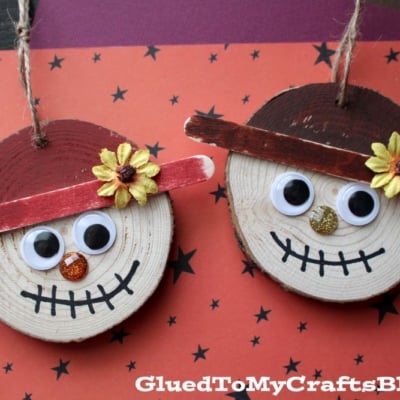 Two scarecrow faces made from round wood slices with googly eyes, stick hats, flowers, and painted smiles make charming wood slice scarecrow ornaments.