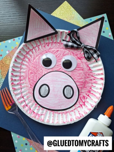 A fork painted pig craft made with a paper plate, colored pink, with paper ears, large eyes, a snout, and a black-and-white bow—surrounded by colored paper, a fork, and glue for creative fun.
