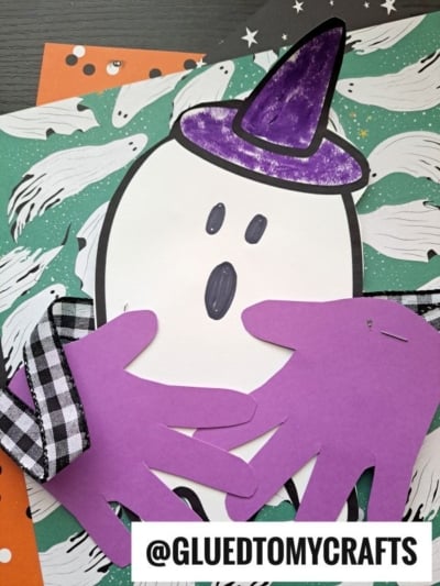 A handprint ghost hug craft featuring a ghost with a purple witch hat and hands made from purple paper, placed on Halloween-themed paper adorned with white ghosts and stars. @GLUEDTOMYCRAFTS text at the bottom.