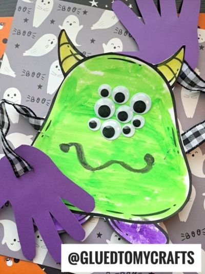A green handprint monster hug craft with googly eyes, a black wavy mouth, yellow horns, and purple paper hands on a Halloween-themed background with ghosts. @GLUEDTOMYCRAFTS is written at the bottom.