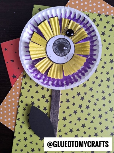 A handmade paper plate Halloween flower craft features a white paper plate bloom with yellow petals, a large eyeball center, a black bug, and a popsicle stick stem, all set against star-patterned paper backgrounds.
