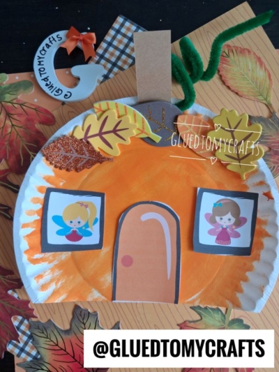 A pumpkin fairy house craft made from a paper plate with fairy stickers as windows, a door, glitter leaves, and pipe cleaner stems, set on fall-themed paper. Text reads @GLUETOMYCRAFTS.