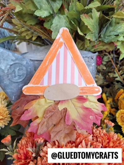 A popsicle stick fall gnome craft featuring a striped orange hat, faux autumn leaves as a beard, and a beige oval nose, surrounded by orange flowers and greenery. A @GLUEDTOMYCRAFTS label is at the bottom.