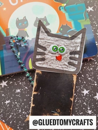 A handmade cat bookmark with googly eyes, a red nose, and a striped face—reminiscent of a playful toilet roll cat—made from paper and a glittery pipe cleaner tail, sits on a book with cartoon cats. @GLUETOMYCRAFTS is written at the bottom.