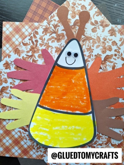 A cute candy corn butterfly craft with a smiling face and googly eyes, decorated with colorful handprint cutout wings, displayed on patterned autumn-themed paper. The text @GLUEDTOMYCRAFTS appears at the bottom.