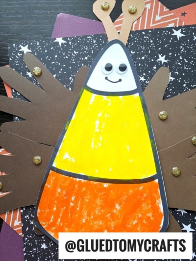 A handmade craft of a candy corn butterfly moth with googly eyes and a smiling face, featuring painted yellow, orange, and white sections. It has brown paper wings with gold glitter spots, set on patterned paper.
