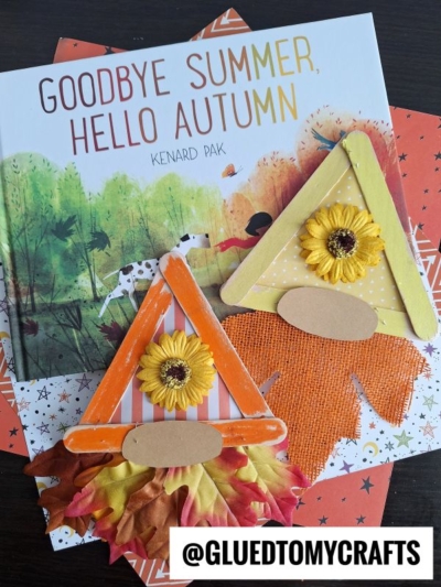 A children’s book titled “Goodbye Summer, Hello Autumn” is displayed with two popsicle stick fall gnome crafts decorated with flowers and leaves, all arranged on top of orange autumn-themed paper.