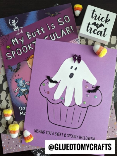 A Halloween craft display with a handprint ghost cupcake card, a “My Butt is So Spooktacular!” book, candy corn, and a “trick or treat” box on patterned paper. @GLUEDTOMYCRAFTS tag is shown.