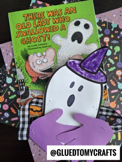 A children’s book titled “There Was an Old Lady Who Swallowed a Ghost!” lies next to Halloween-themed paper and a cute handprint ghost hug craft with a purple hat and hands. A label at the bottom reads @GLUEDTOMYCRAFTS.