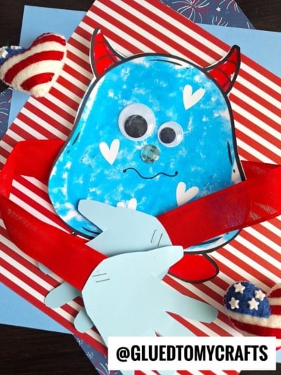 A handmade blue handprint monster hug craft with googly eyes, red horns, paper heart shapes, and two paper hands on a striped background. Decorated with patriotic heart plushies. Text reads @GLUEDTOMYCRAFTS.