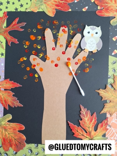 A q-tip painted fall tree craft made from brown paper, decorated with orange and yellow dots, features a small owl cutout perched on one of the fingers. Fall leaves and a Q-tip are arranged on a black background to complete this charming autumn scene.