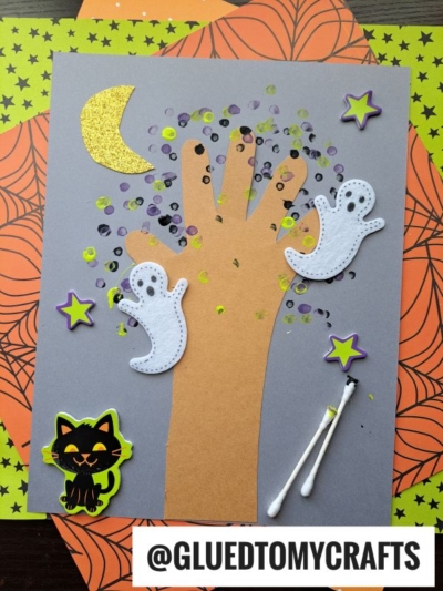 A Halloween craft features a q-tip painted fall tree made from a handprint on gray paper, decorated with ghost and star stickers, a cat sticker, bright painted dots, two cotton swabs, and a yellow moon. Background papers display spooky spiderweb and star patterns.