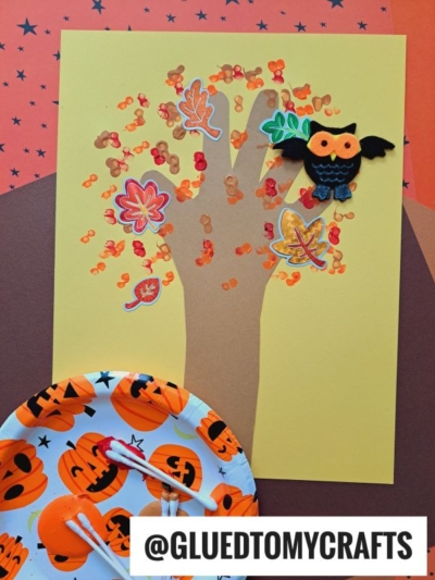 A fall craft featuring a q-tip painted fall tree with dot-painted leaves, colorful leaf stickers, and a felt owl with bat wings on a yellow background. Supplies and orange paint are on a pumpkin-themed plate nearby.