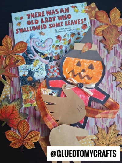 A children’s book titled There Was an Old Lady Who Swallowed Some Leaves! lies on a fall-themed surface with paper leaf cutouts. Next to it, a handmade pumpkin-headed scarecrow extends its arms as if ready for a warm scarecrow hug.