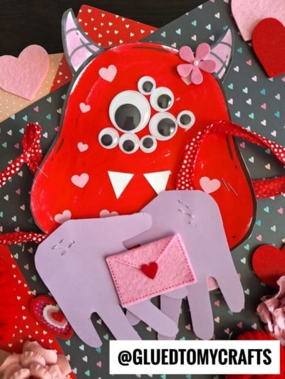 A red, heart-themed handprint monster hug craft with googly eyes, horns, and paper hands holding a pink envelope, surrounded by paper hearts. The @GLUEDTOMYCRAFTS watermark is at the bottom.
