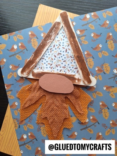 A fall-themed craft features a popsicle stick fall gnome created from popsicle sticks, a fabric leaf, and patterned paper, all arranged on blue paper with an autumn bird print. @GLUEDTOMYCRAFTS is written at the bottom.