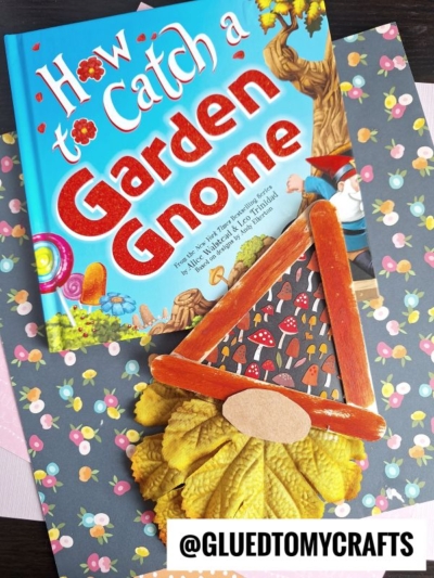A colorful book titled How to Catch a Garden Gnome sits on floral paper, accompanied by a handmade popsicle stick fall gnome crafted from leaves, a red triangle, and a popsicle stick. The handle @GLUEDTOMYCRAFTS is at the bottom.