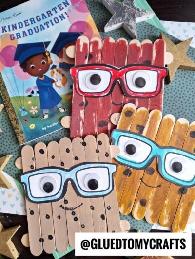 Three Popsicle Stick Smart Cookie crafts, painted as faces with large googly eyes and glasses, are displayed next to a Kindergarten Graduation! book and gold stars on a polka dot background.