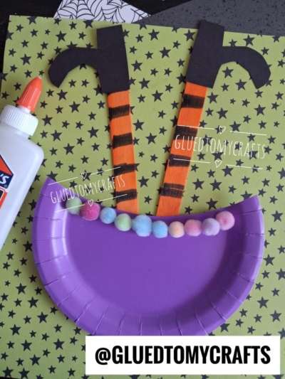 A purple paper plate with colorful pom-poms, two striped witch legs made of paper, and a bottle of glue are all arranged on a green star-patterned background, making this the perfect setup for a fun witch cauldron art project.