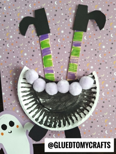 A paper plate monster craft with a black-painted mouth, purple pom-poms for teeth, striped antennae, and black paper shapes on a purple starry background. A cute ghost cutout adds charm—perfect inspiration alongside witch cauldron art! Text: @GLUEDTOMYCRAFTS.