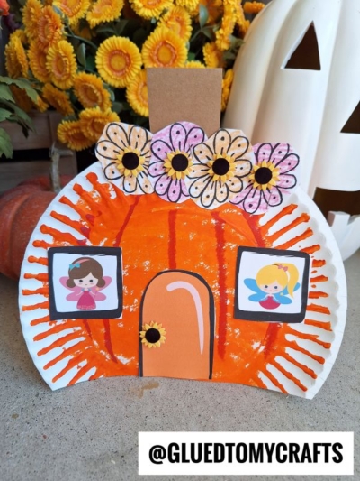 Paper plate pumpkin craft decorated to look like a charming pumpkin fairy house, with flowers and cartoon fairy pictures as whimsical windows, plus a brown stem, set in front of yellow flowers and a white pumpkin. @GLUETOMYCRAFTS text is at the bottom of the image.