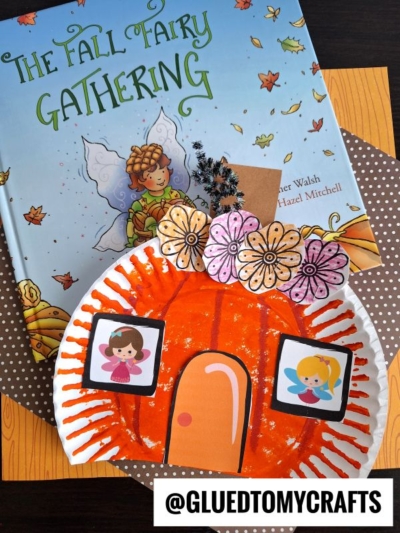 A childrens book titled The Fall Fairy Gathering sits behind a paper plate craft decorated like a pumpkin fairy house with drawn flowers and two fairy illustrations. The @gluedtomycrafts handle is visible at the bottom.