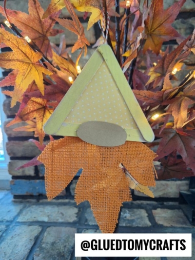 A popsicle stick fall gnome, crafted from a yellow triangle, paper nose, and orange fabric leaves for a whimsical beard, hangs among vibrant autumn leaves. @GLUEDTOMYCRAFTS.