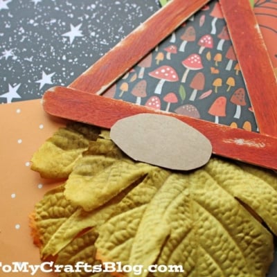 A popsicle stick fall gnome craft featuring orange popsicle sticks as a triangle hat, yellow faux leaves for a beard, and a small oval paper nose, displayed on autumn-themed paper backgrounds.