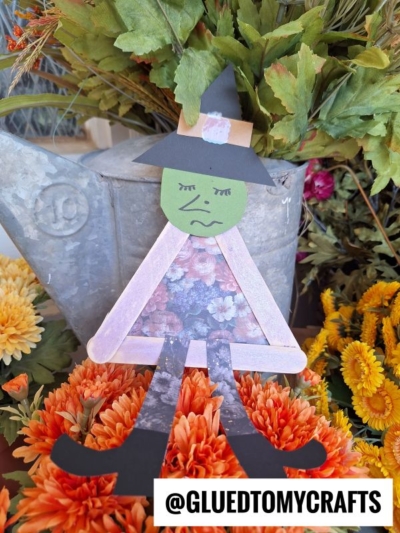A popsicle stick Halloween witch with a green face, black hat, and sad expression sits among colorful artificial flowers. Popsicle stick arms and legs complete this charming craft. Text at the bottom reads @GLUEDTOMYCRAFTS.